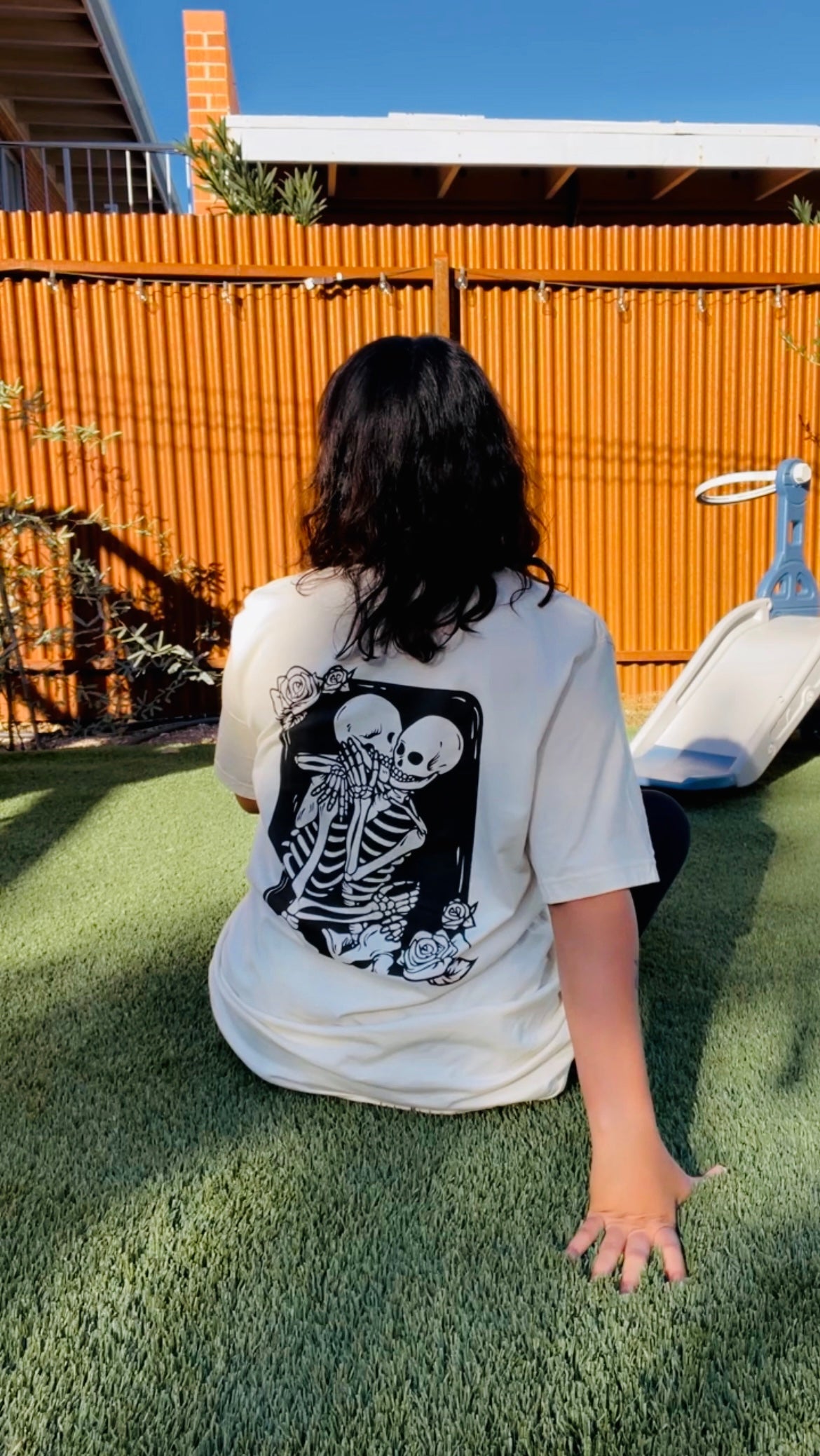 A person seated on the lawn donning the 'Amor Eterno Unisex Tee' featuring a skeleton design